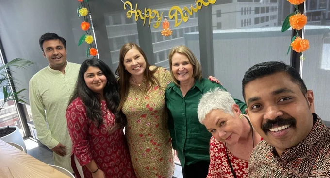 A cheerful group of colleagues dressed in festive attire pose together under a "Happy Diwali" banner, celebrating the festival in a decorated office space