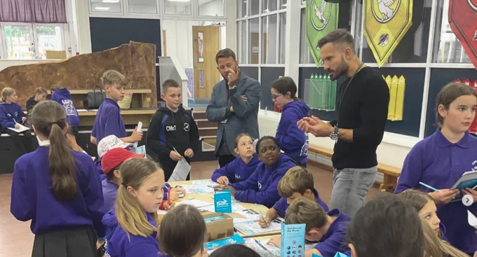 Group of schoolchildren in purple leavers hoodies sitting around tables with activities, guided by adults in a school hall