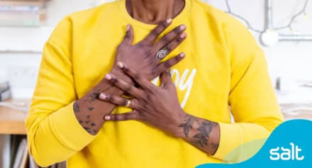 A person in a yellow sweatshirt and tattooed arms, holds their hands over their heart in a warm, showing confidence
