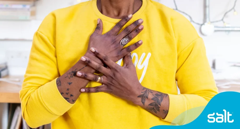 A person in a yellow sweatshirt and tattooed arms, holds their hands over their heart in a warm, showing confidence