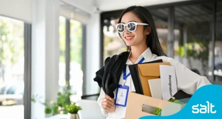 Woman wearing sunglasses, smiling and carrying a box with folders and a paper labeled “resignation”, symbolising a positive career transition