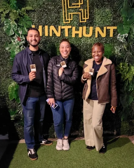 Three people smiling and holding cards in front of a HintHunt backdrop