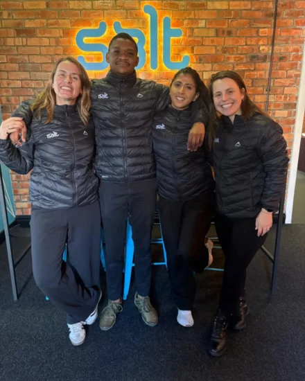 Four smiling team members wearing matching Salt-branded black jackets pose together in front of a brick wall with a glowing Salt logo