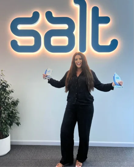 Smiling woman holding Salt awards in both hands, standing in front of a large illuminated Salt logo at the office