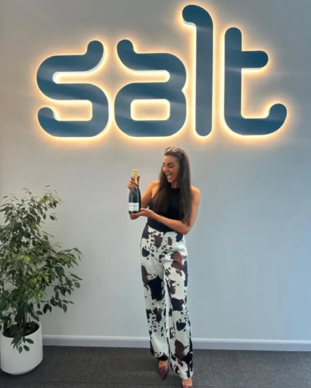 Smiling woman holding a champagne bottle in front of a glowing Salt logo at the office, celebrating a promotion