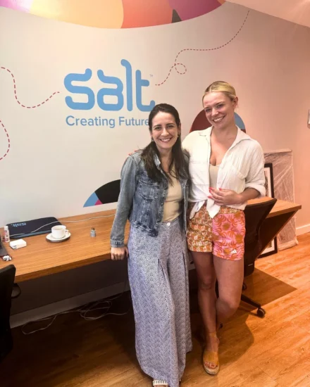 Two women smiling and standing in front of a "Salt Creating Futures" wall inside an office space