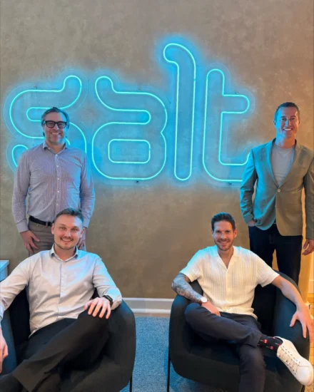 A group of four Salt employees posing in front of a glowing "Salt" sign at an office, with two sitting in chairs and two standing behind them