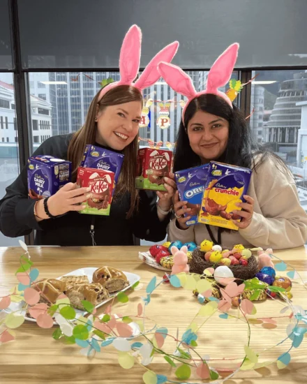 Two women wearing pink bunny ears hold Easter chocolates and smile at the camera, with festive treats and decorations on the table in front of them